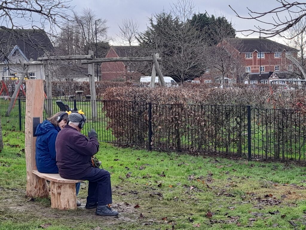 An older couple sit on a carved wooden bench in front of Gainsborough Square playground. They have headphones on.