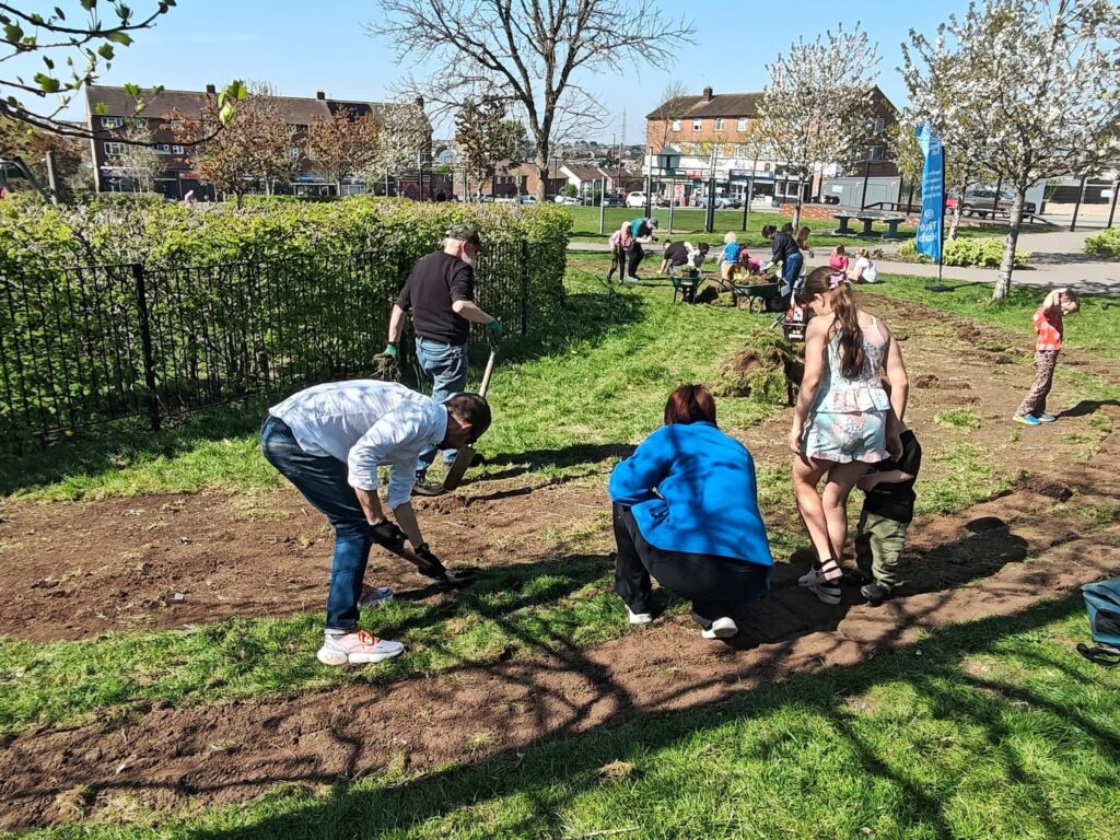 Description: 15 people of all ages use rakes, wheelbarrows and pick up turf. It's a sunny day and they are on Gainsborough Square with bushes, shops and blossoming trees and an LNT flag behind them.