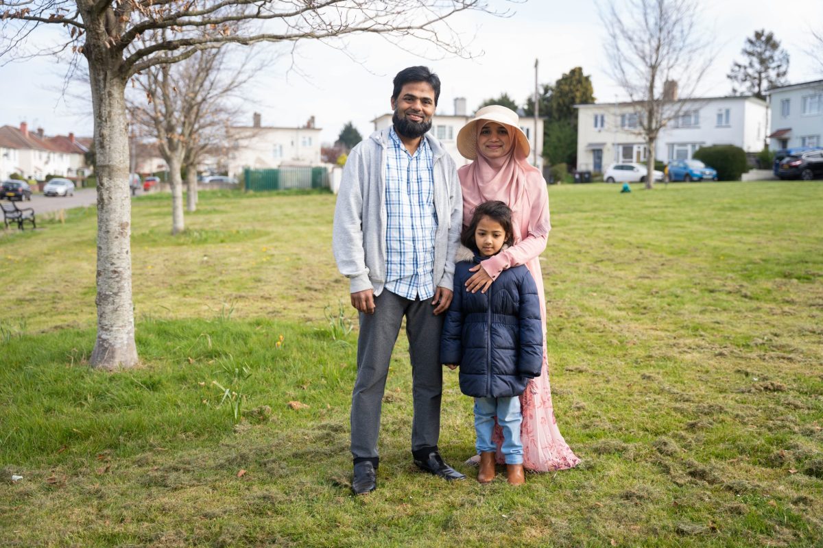 Huma, Noman and their young daughter stand smiling on a housing green with trees