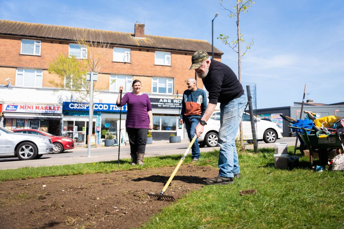 An older man rakes a bare patch of land in front of some shops on a sunny day.