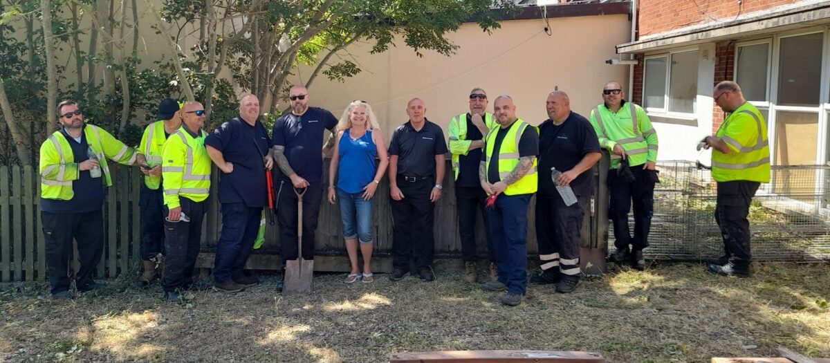 11 volunteers in Bauer Outdoor Media t-shirts and hi vis line up along a fence. In the middle is Maria from LNT, a white woman with blonde hair and a blue LNT vest. 