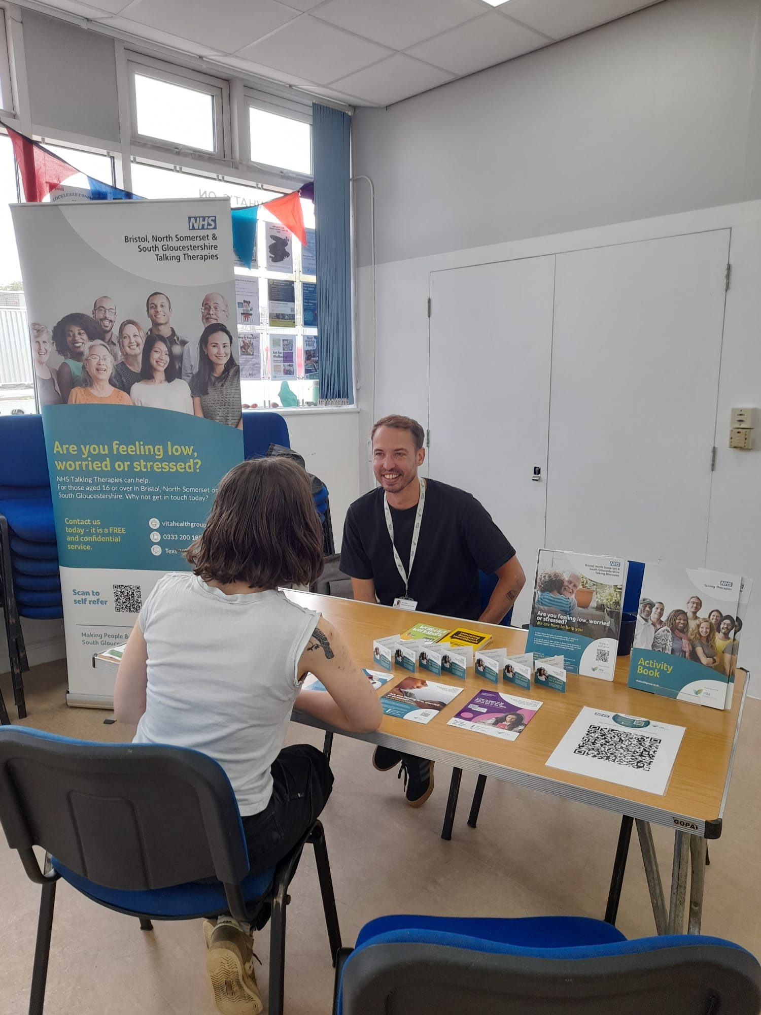 A young woman sits at a table in front of a man. There is a banner and lots of leaflets for NHS talking therapies.