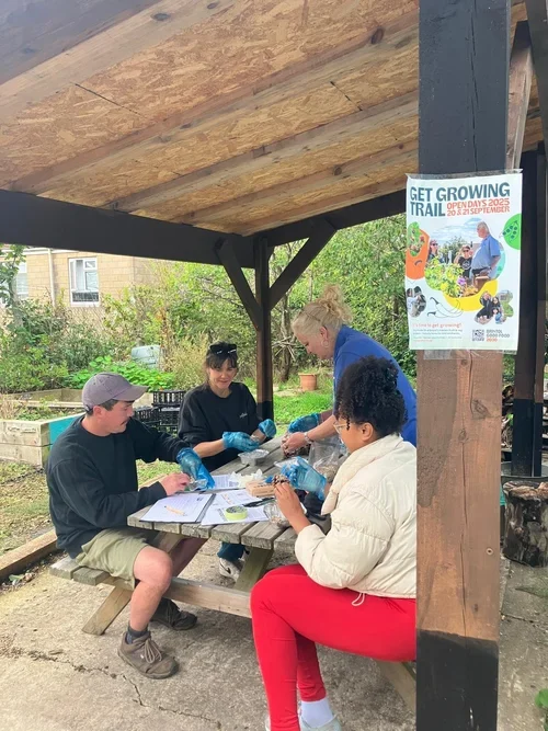 Four adults make birdfeeders at an outdoor table. In front is a post with a get growing trail poster and behind are raised beds