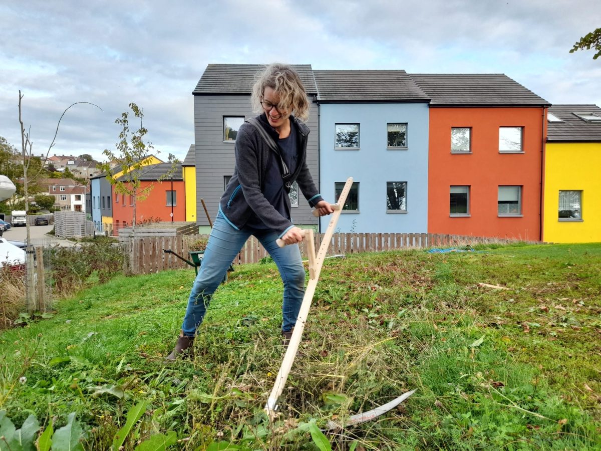 A woman uses a scythe to cut grass in front of colourful houses.
