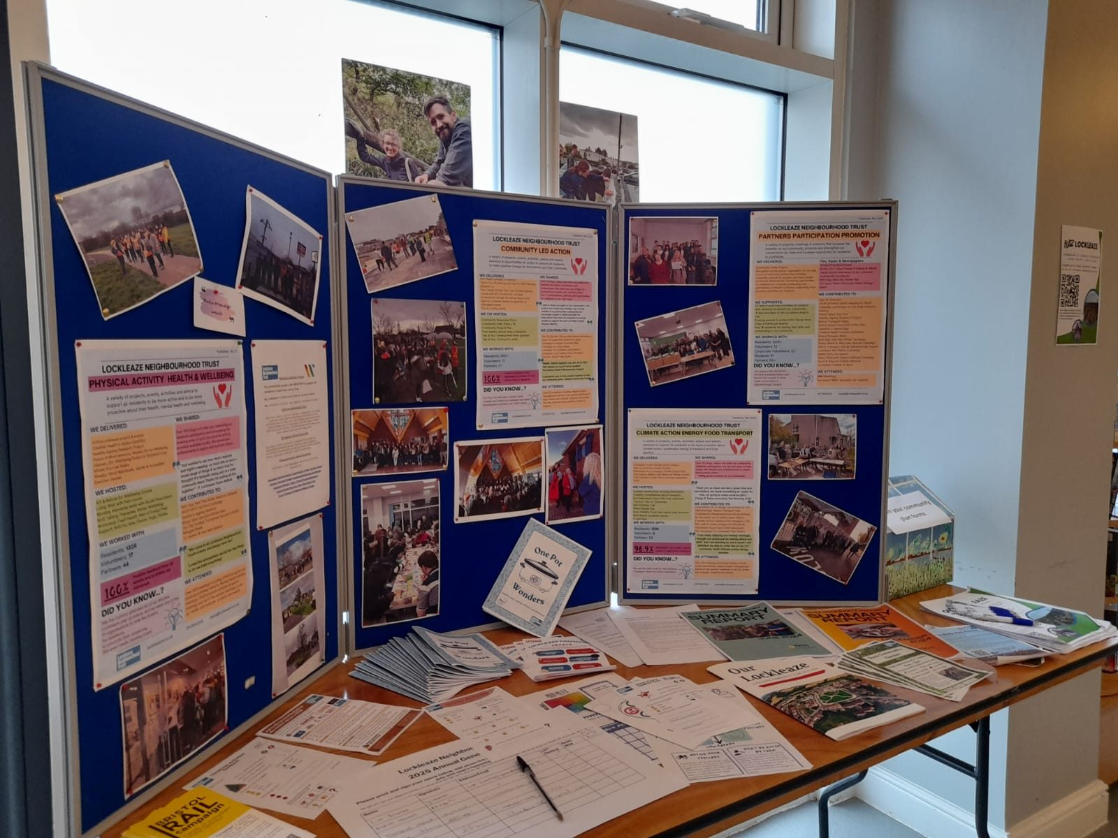 A blue vertical display board on a table with sign in sheets, a 2018-24 community plan, recipe books and pictures of events from across the year