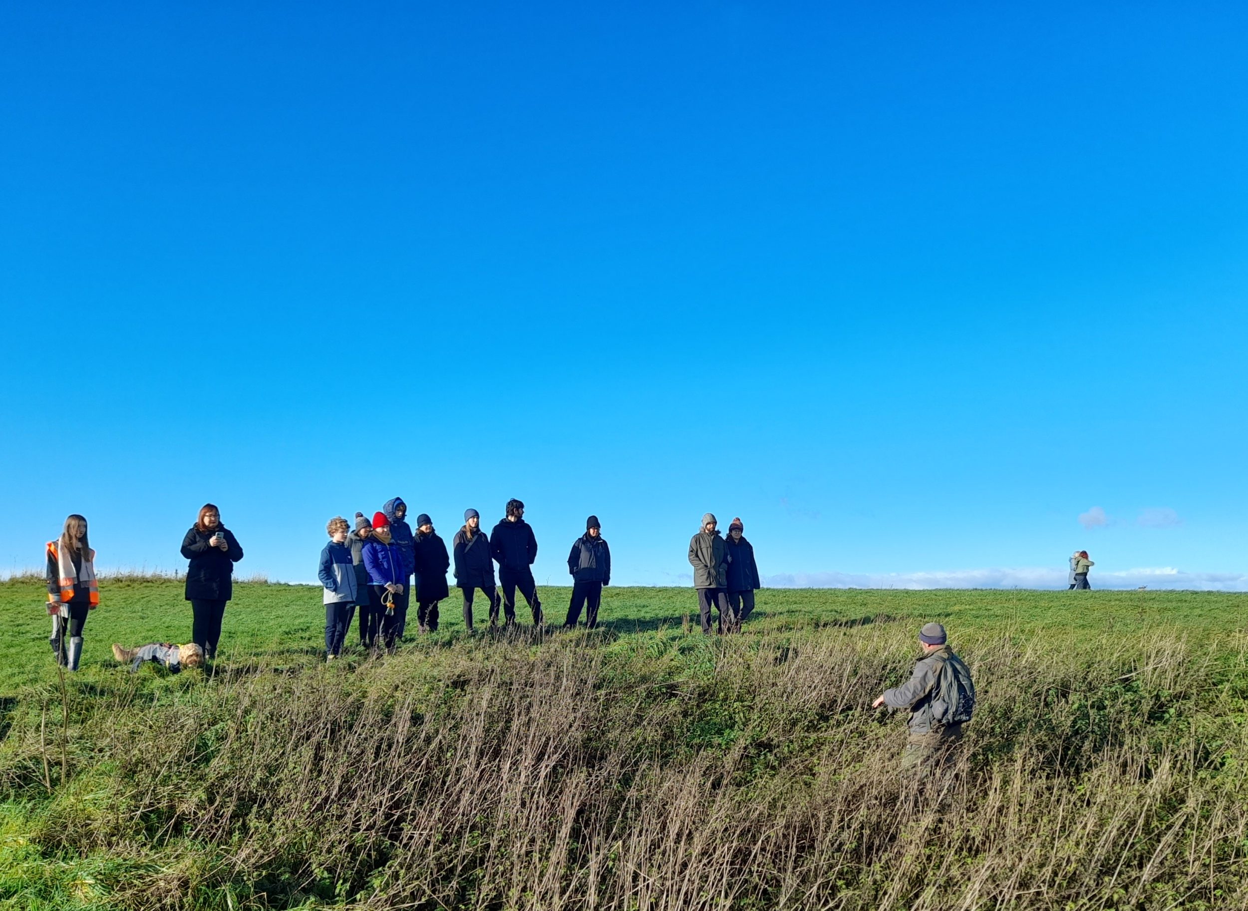 steve, wearing green outdoor gear is in a patch of long grass. above him a group of people listen