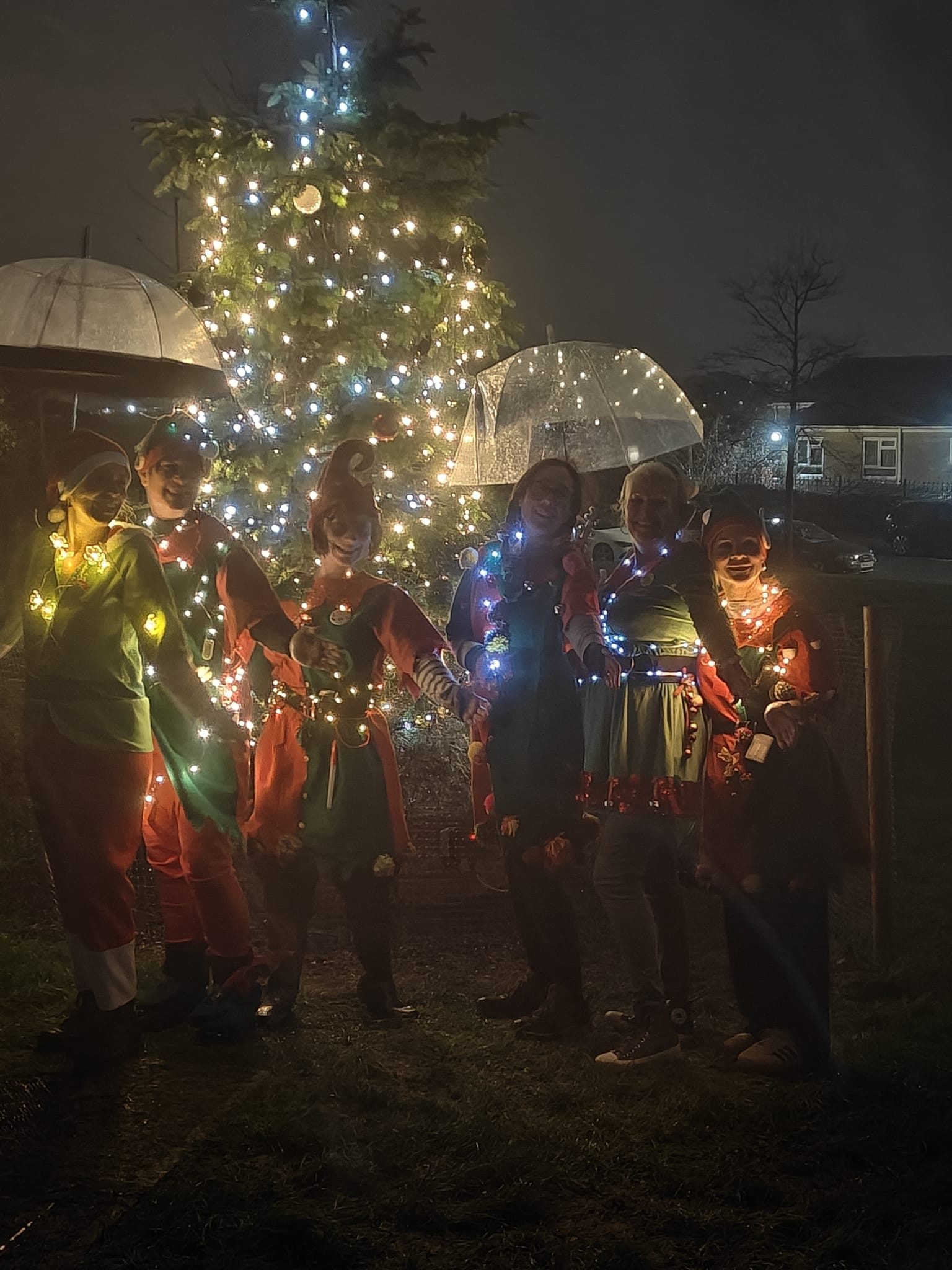 six women dressed as elves with fairy lights on them stand in front of a brightly lit up christmas tree in the dark