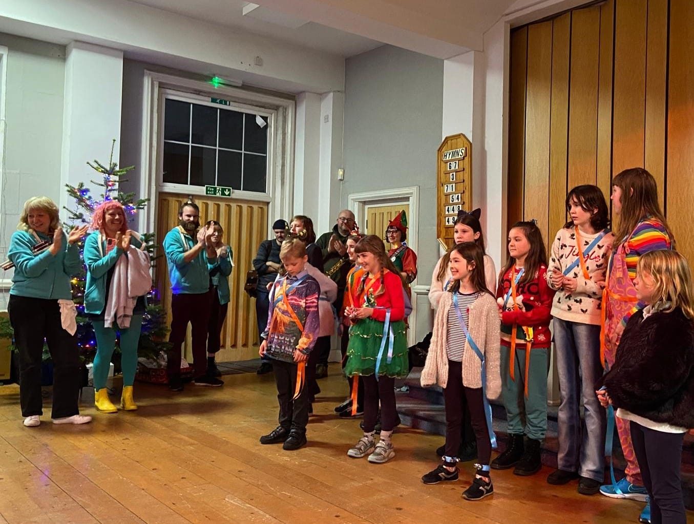 a group of children stand smiling wearing blue and orange ribbons as people applaud from the side