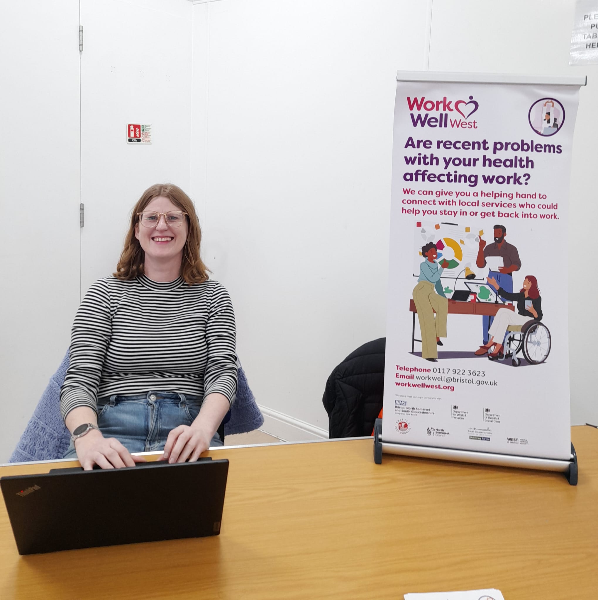 a woman smiles at her laptop at a table with a banner for work well west, printed with 'are recent problems with your health affecting work?'
