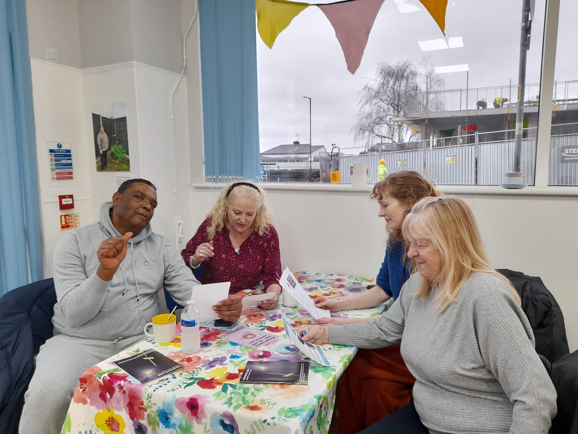 A Black man and three white women chat at a table with mugs of tea and useful leaflets
