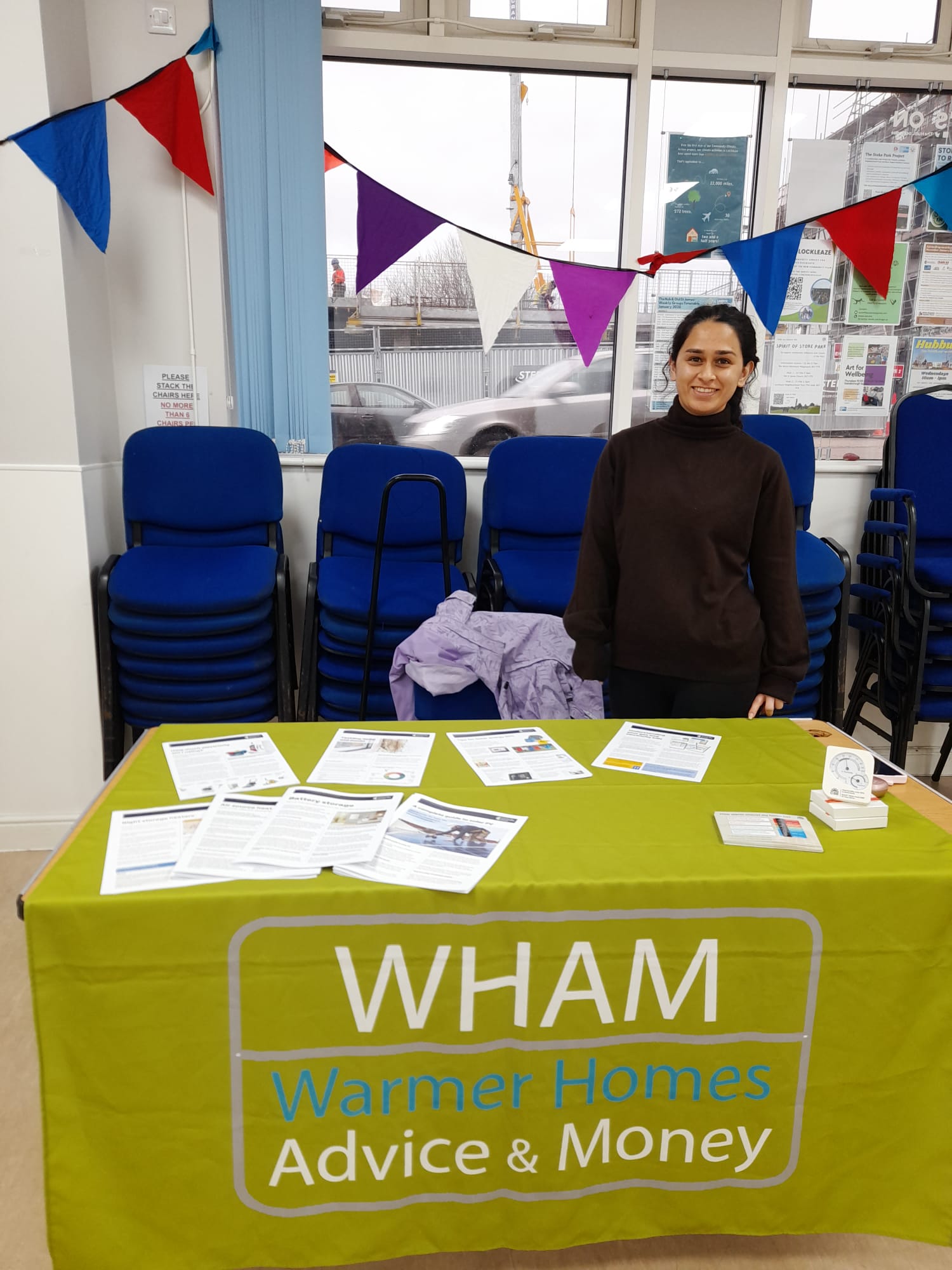 a young Asian woman stands behind a desk with printed materials about home energy efficiency, with a blanket saying 'WHAM: warmer homes advice & money'
