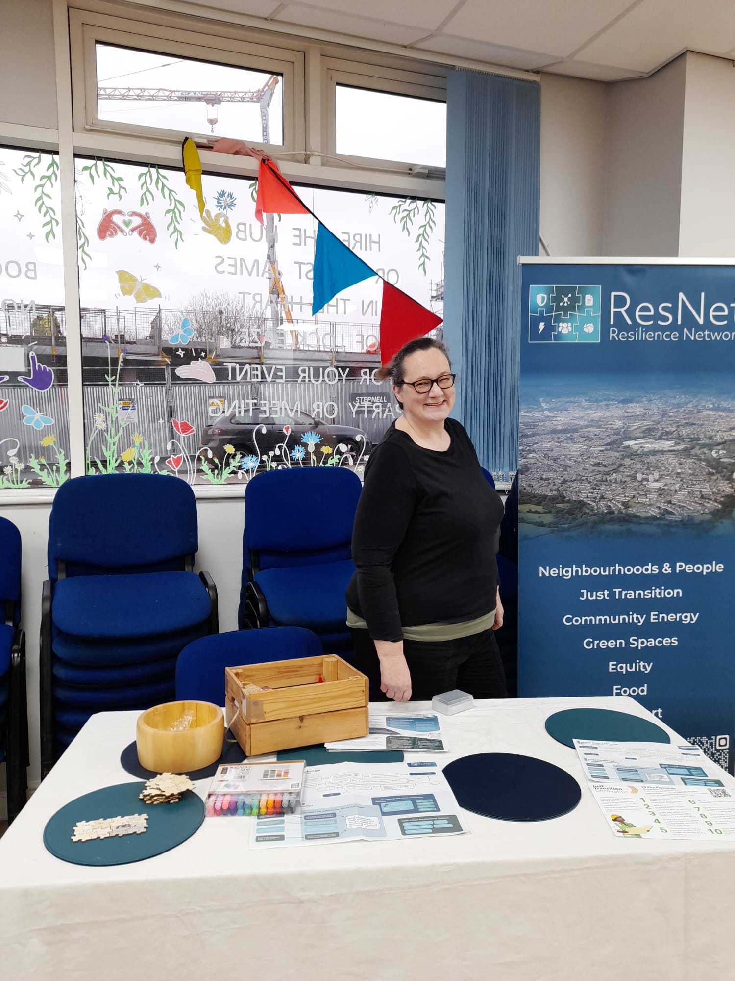 a white woman smiles behind a desk with materials for resnet, which reads 'resilience network, neighbourhoods and people, community energy, just transition' etc