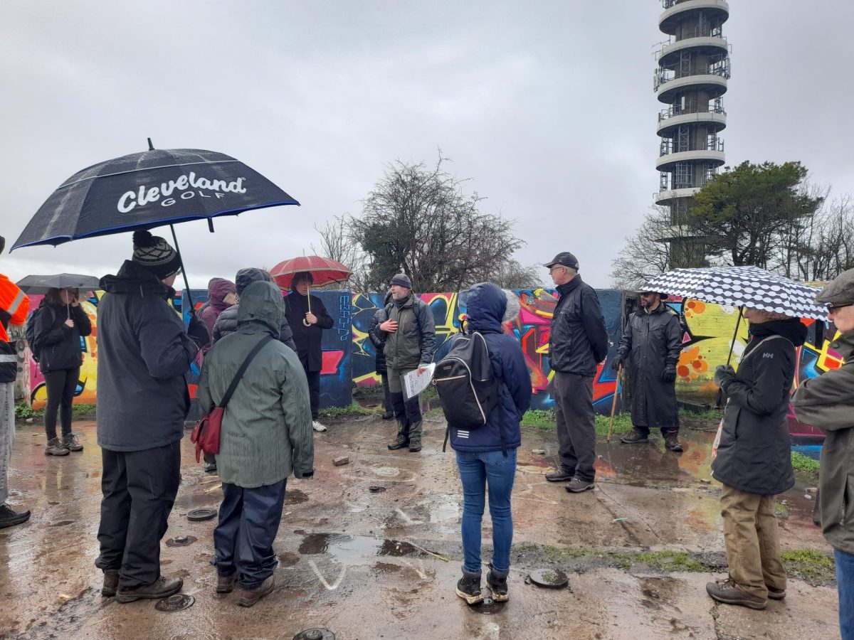 twenty people with umbrellas gather in front of brightly graffitid concrete gun battery structures