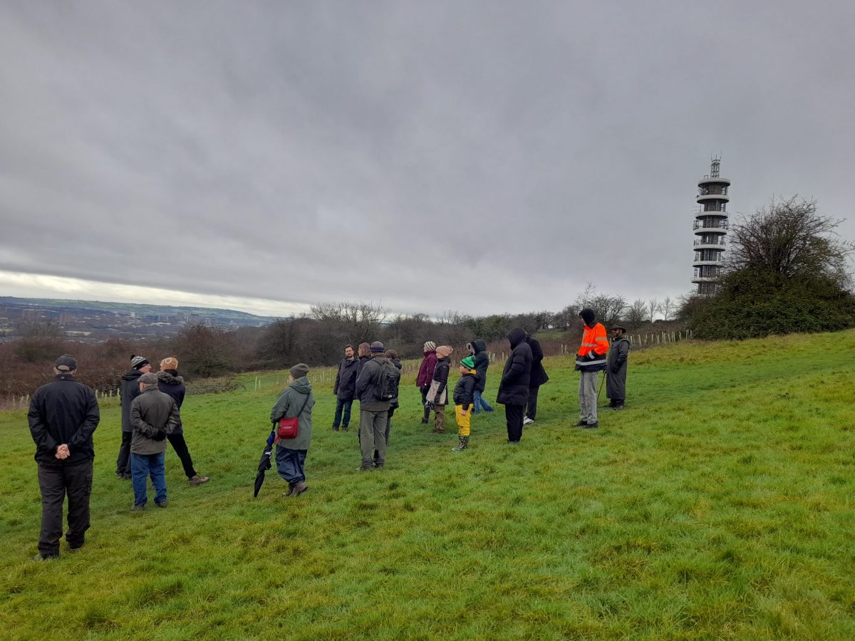 twenty people of all different ages walk up a grassy slope to the purdown tower on a grey day.