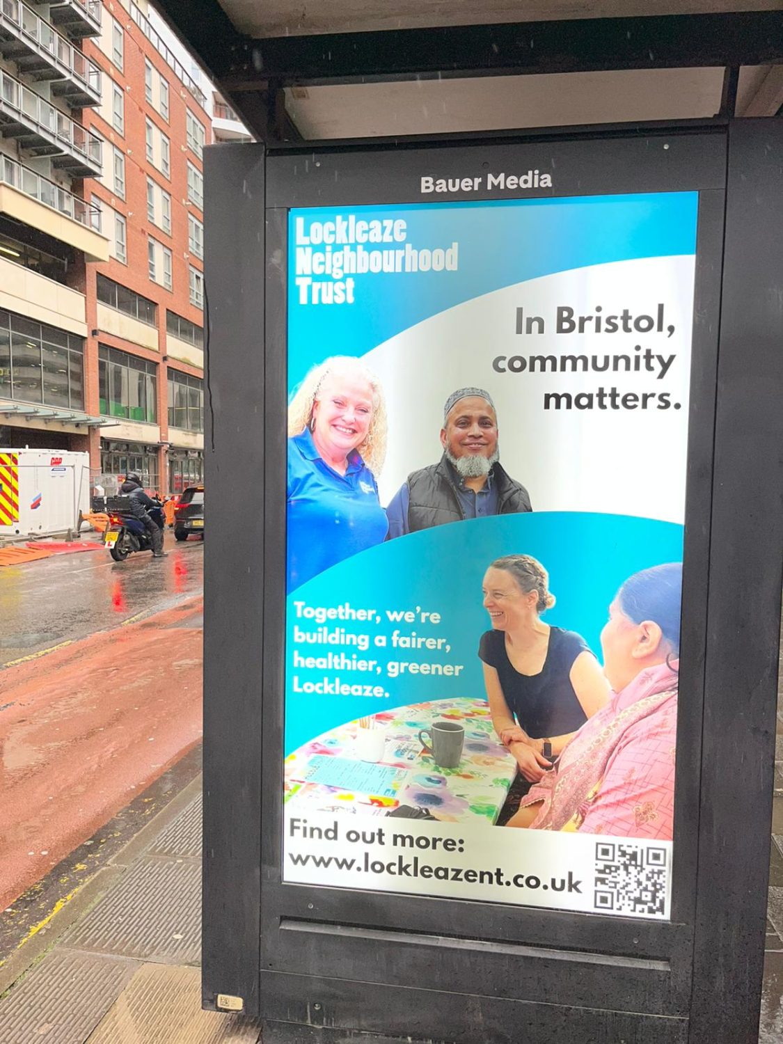 a bus stop advert about the height of a person. it features four smiling people, one in an LNT shirt, and the words 'Lockleaze Neighbourhood Trust. In Bristol, Community Matters. Together, we're working for a fairer, healthier, greener Lockleaze. Find out more: www.lockleazent.co.uk'