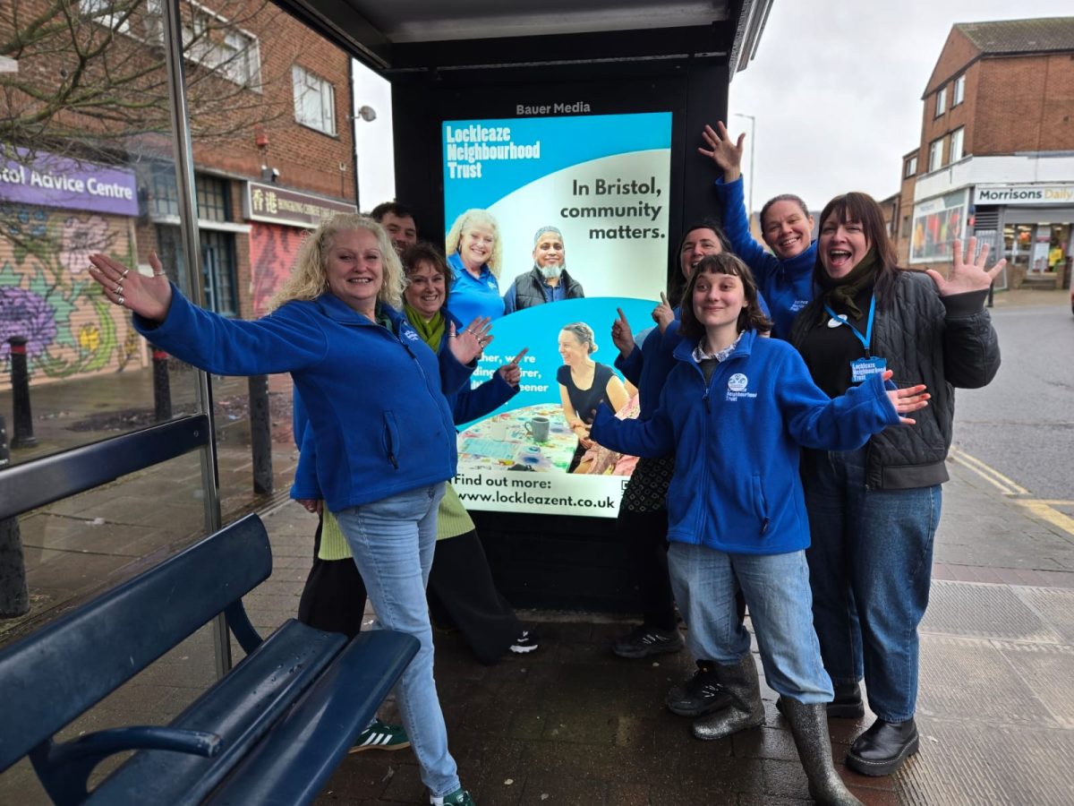 seven people stand in a bus shelter, smiling and gesturing to a bus ad featuring lockleaze neighbourhood trust logo, smiling people and the statement 'in Bristol, community matters'.