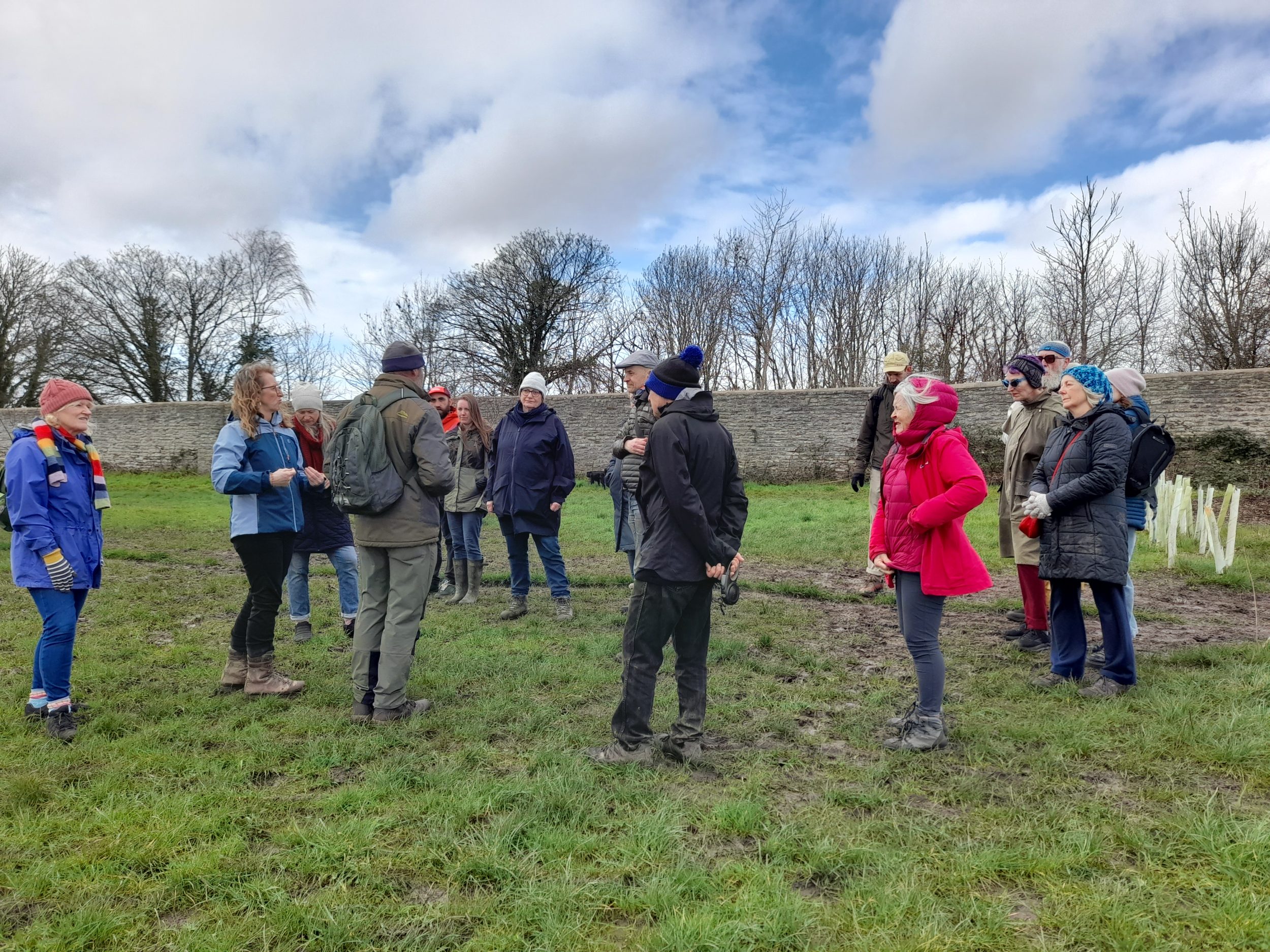 a group of walkers in colourful winter clothes pause on a muddy path through a field. there are new trees in green plastic tree guards behind