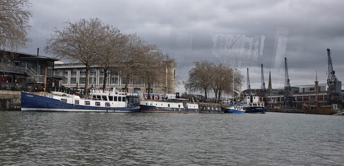looking over grey ruffled water at ships and tall buildings