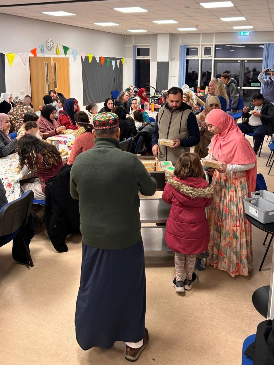a young woman in a pink hijab serves food from a trolley to two men and a little girl. Behind are multiple tables of people eating, in a well-lit community hall with grey drapes and bunting