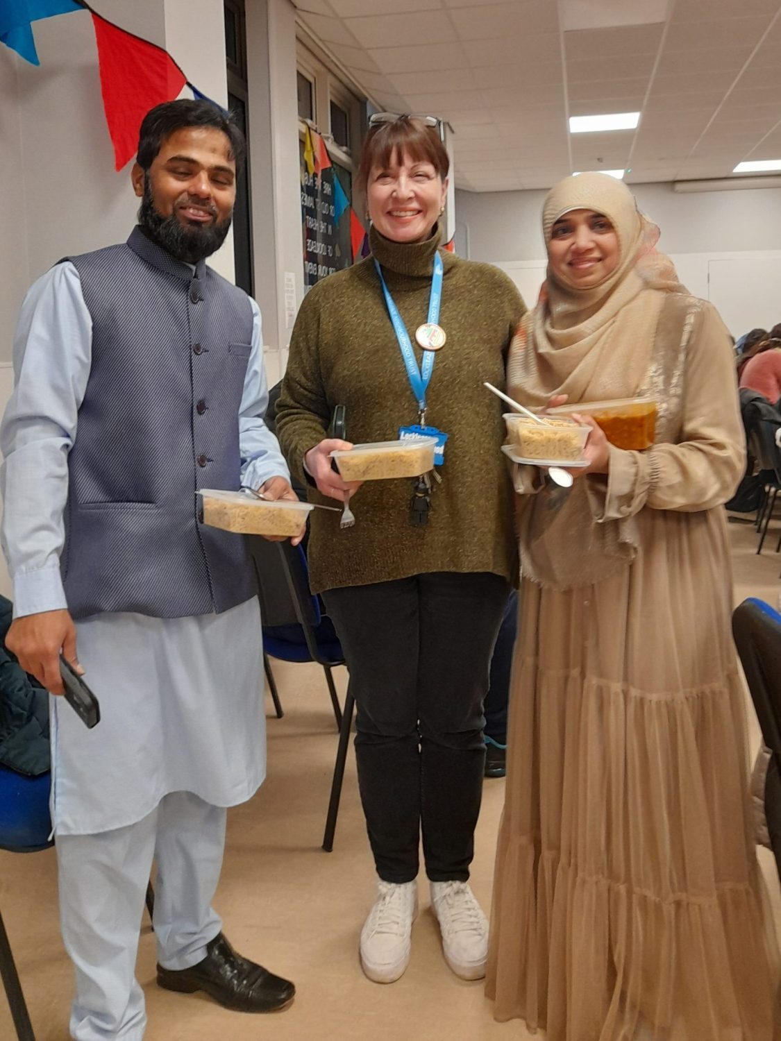 a Muslim couple in celebratory clothes and white woman in LNT lanyard smile with tubs of food