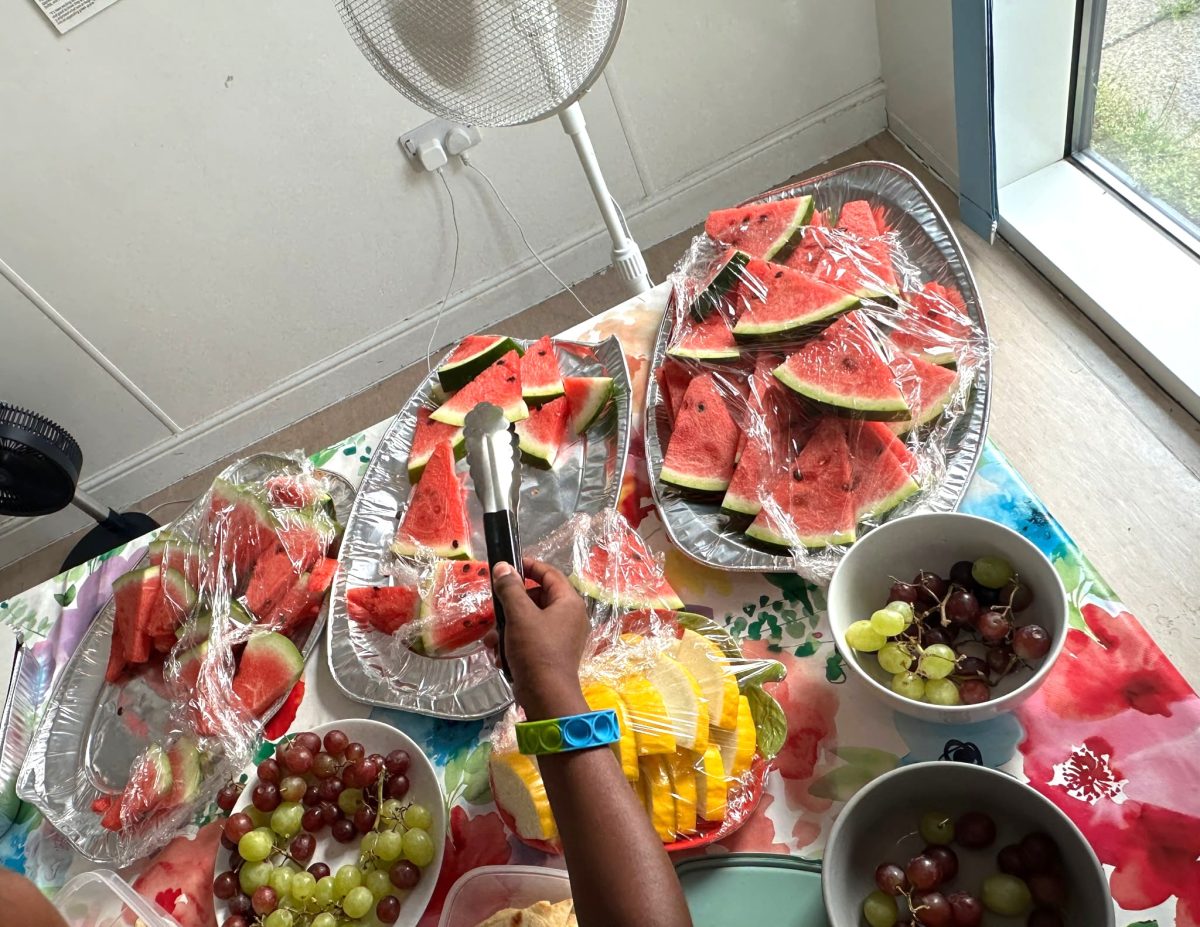 a table with slices of watermelon, yellow melon and grapes. a Black person's hand holds a pair of tongs over the melon