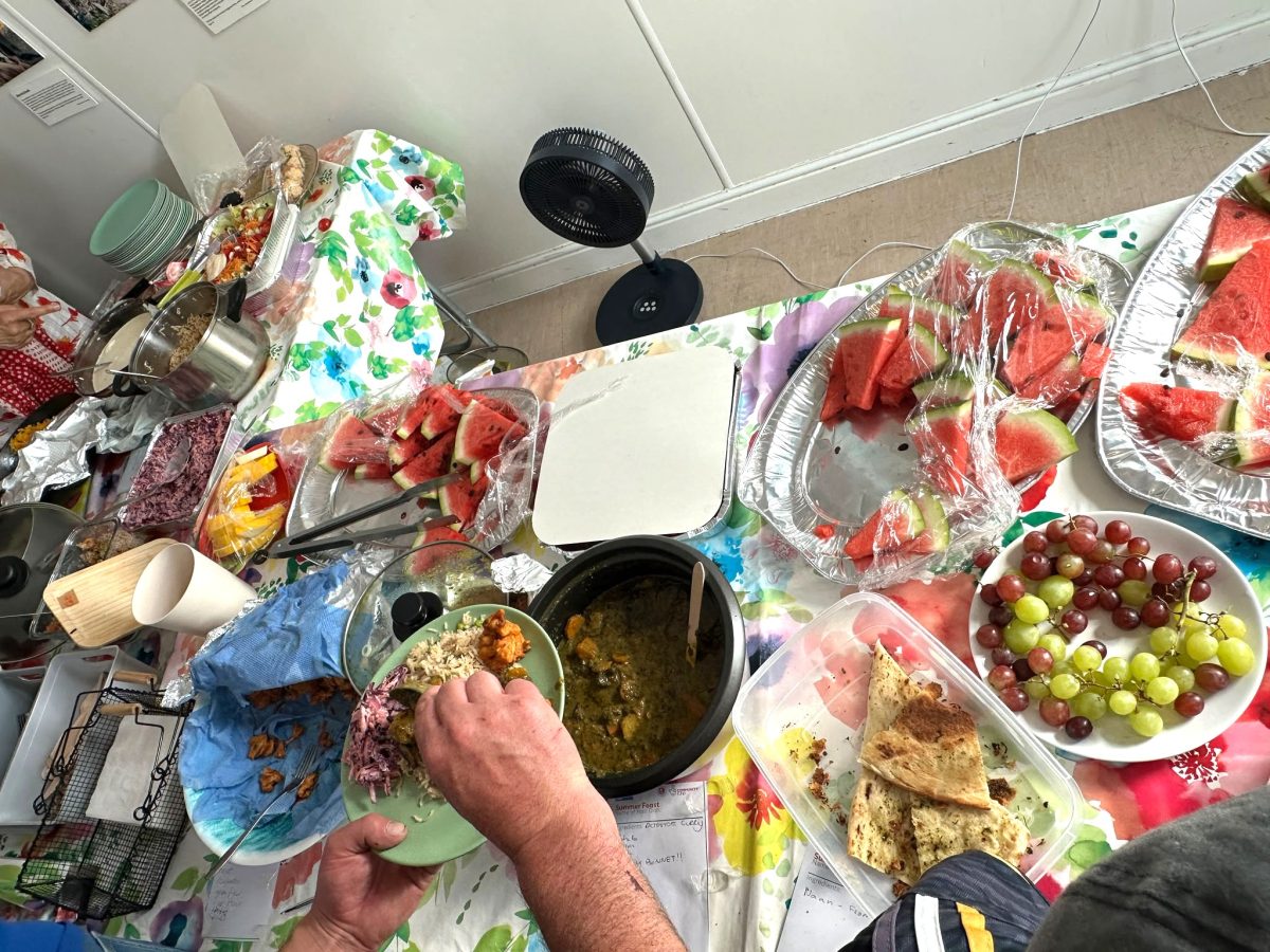 watermelon, grapes, flatbread, multiple salads, curry and rice on a table with a flowery cloth. an older white person's hand is shown choosing food.