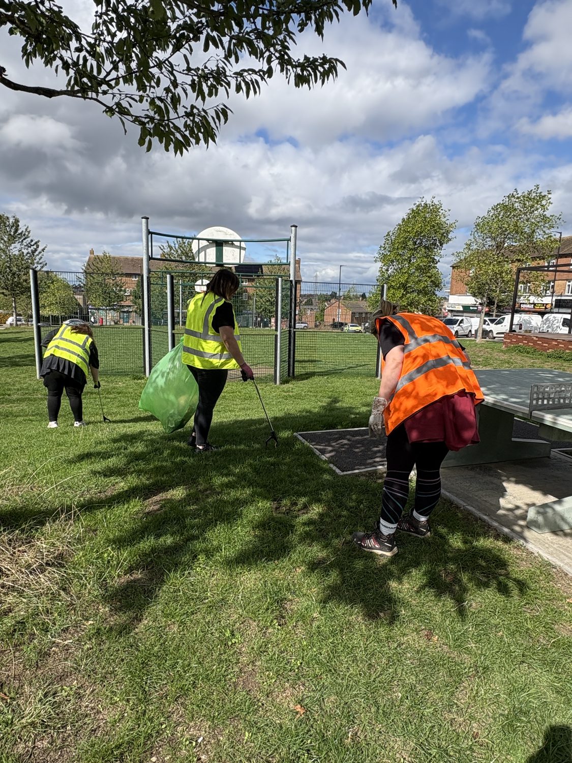 three women in high vis vests use litterpickers on a grassed public sports pitch