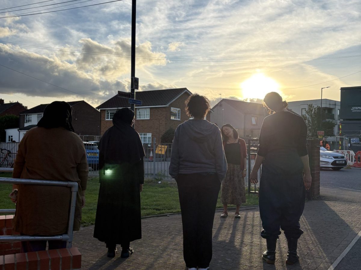 a group outside stand facing Naomi, each tilting their head to the side. Behind the sun is setting with yellow rays in a blue sky.