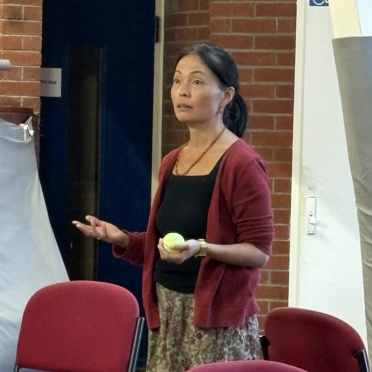 a slim East Asian woman in her early fifties speaks with her hands upheld, one holding a tennis ball