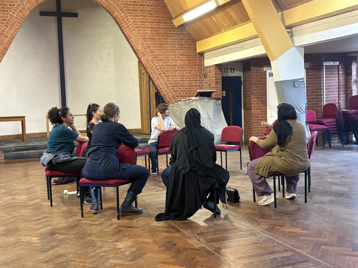 five women and a man, of various ethnicities, sit facing in in a circle in a church hall