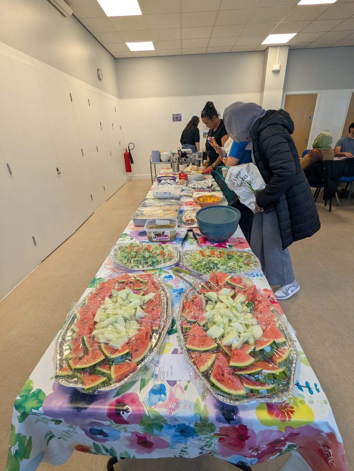 three people lean over a very long table with multiple dishes including cut fruit and a flowery tablecloth